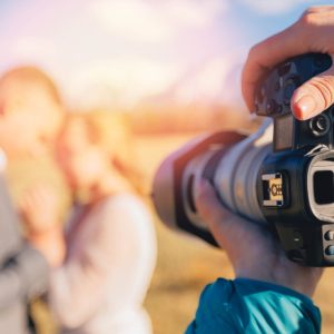 Una forocamera inquadra una coppia sfocata in un ambiente esterno, con qualcuno che regola la telecamera in primo piano, come descritto in "Fotografia di Eventi: Tecniche per Documentare Eventi e Celebrazioni.