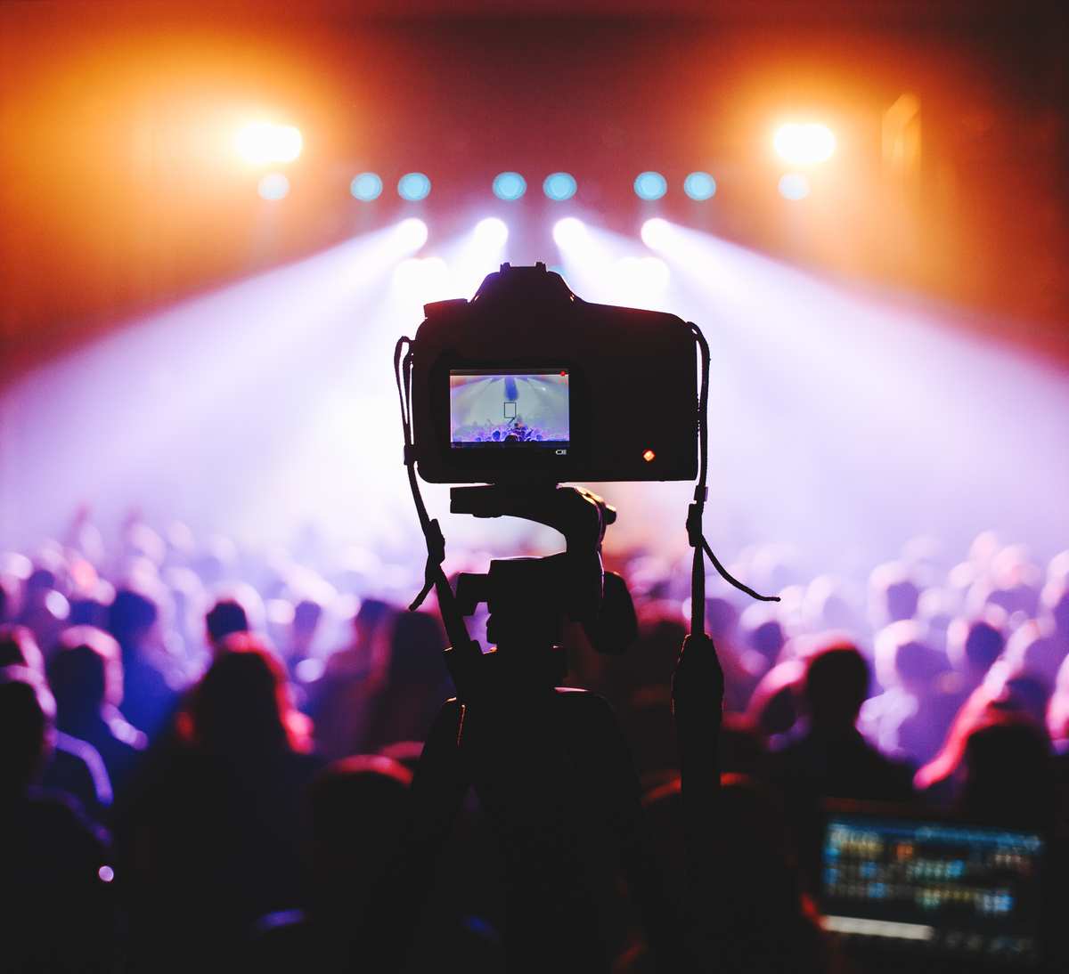 Una Telecamera Montata Su Un Treppiede Cattura Riprese Video Di Una Folla A Un Concerto, Con Luci Intense Del Palco Che Illuminano La Scena.